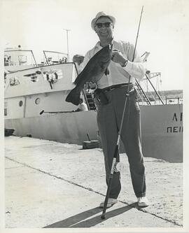 Hombre extranjero sosteniendo pez junto a caña de pescar y barco de fondo.