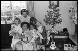 Casa de la familia López Arango, Canal 6 TV. Navidades. Mujer posando junto a cuatro niñas que llevan juguetes en sus brazos. Están ubicadas en el sillón de la sala de la casa, junto a decoración navideña.